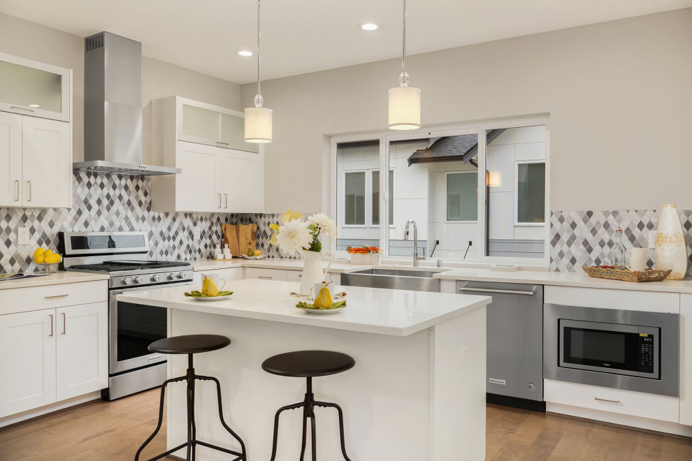 Design ideas for a transitional l-shaped kitchen in Seattle with a farmhouse sink, shaker cabinets, white cabinets, multi-coloured splashback, stainless steel appliances, medium hardwood floors, with island, brown floor and white benchtop.