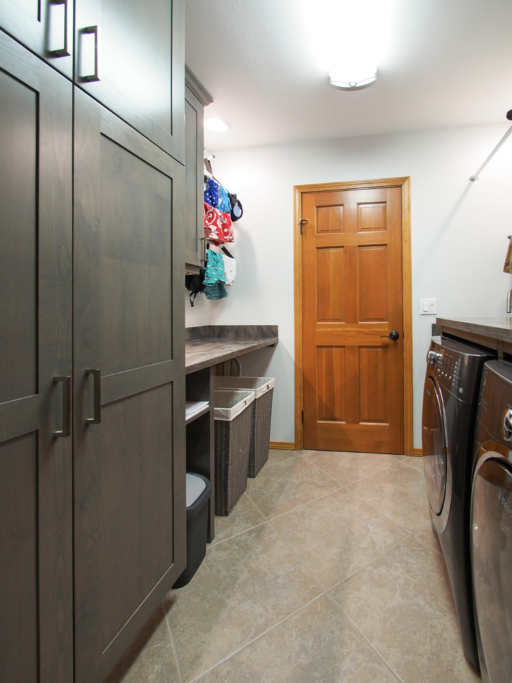 Photo of a mid-sized transitional galley dedicated laundry room in Seattle with an undermount sink, recessed-panel cabinets, dark wood cabinets, solid surface benchtops, blue walls, porcelain floors, a side-by-side washer and dryer, brown splashback, beige floor and brown benchtop.