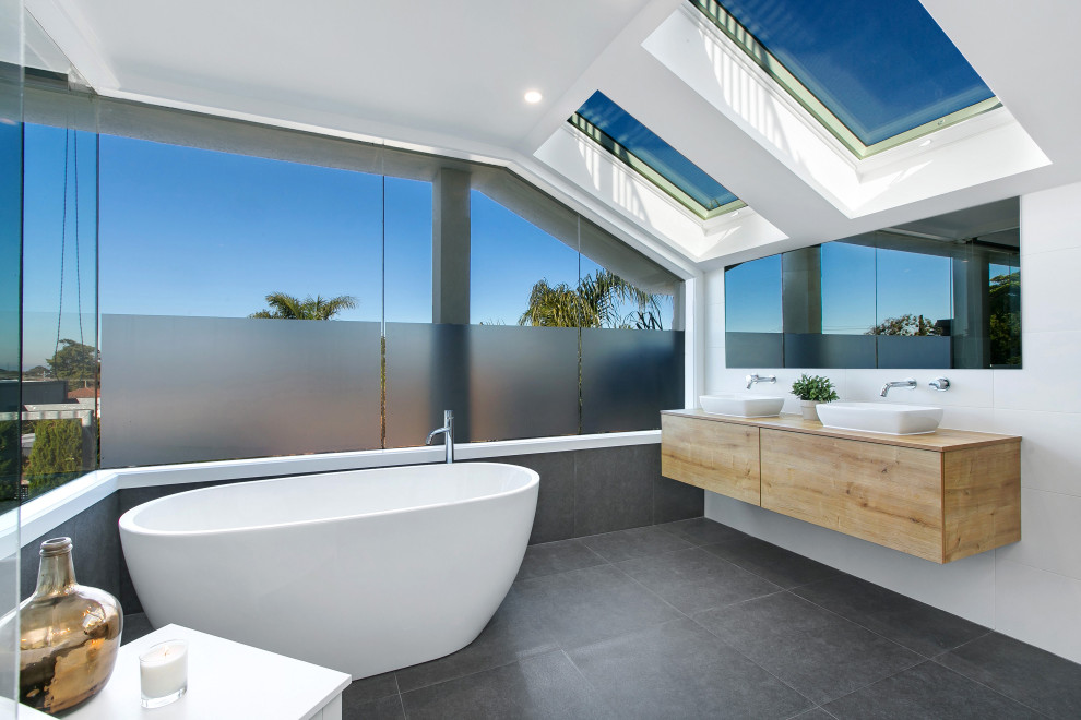 Modernes Badezimmer mit hellen Holzschränken, freistehender Badewanne, weißen Fliesen, weißer Wandfarbe, Keramikboden, Waschtisch aus Holz, grauem Boden, Doppelwaschbecken und schwebendem Waschtisch in Melbourne