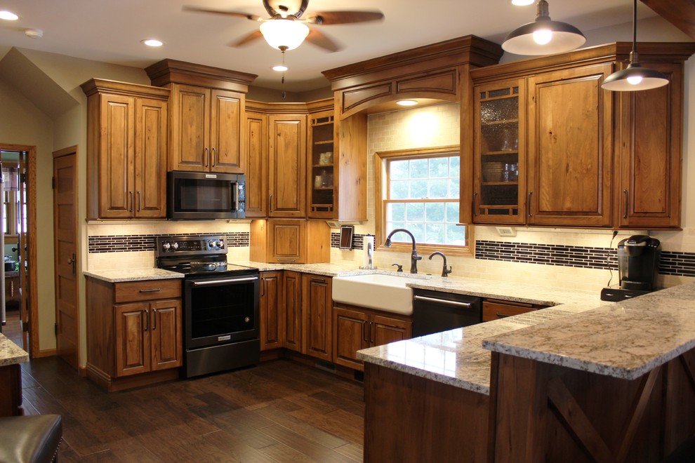 Photo of a large country u-shaped eat-in kitchen in Other with a farmhouse sink, raised-panel cabinets, medium wood cabinets, granite benchtops, beige splashback, travertine splashback, black appliances, medium hardwood floors, a peninsula, brown floor and beige benchtop.