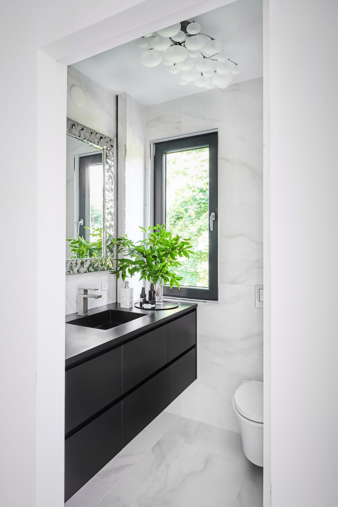 Идея дизайна: туалет среднего размера в современном стиле с плоскими фасадами, черными фасадами, инсталляцией, белой плиткой, керамогранитной плиткой, белыми стенами, полом из керамогранита, монолитной раковиной, столешницей из искусственного камня, белым полом, черной столешницей и подвесной тумбой