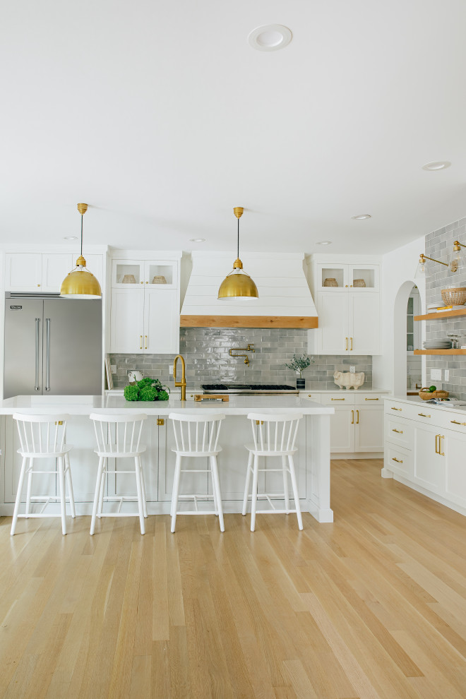 Design ideas for a large nautical l-shaped open plan kitchen in Other with a belfast sink, shaker cabinets, white cabinets, engineered stone countertops, grey splashback, metro tiled splashback, stainless steel appliances, light hardwood flooring, an island, brown floors and white worktops.