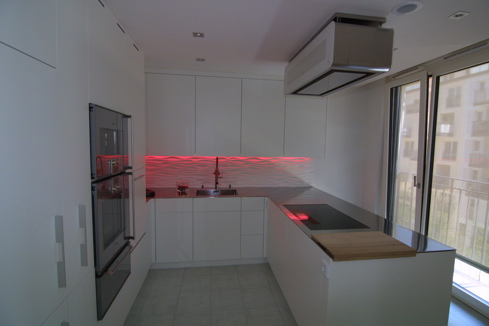This is an example of a contemporary u-shaped open plan kitchen in Munich with an integrated sink, flat-panel cabinets, white cabinets, stainless steel worktops, white splashback, stainless steel appliances and a breakfast bar.