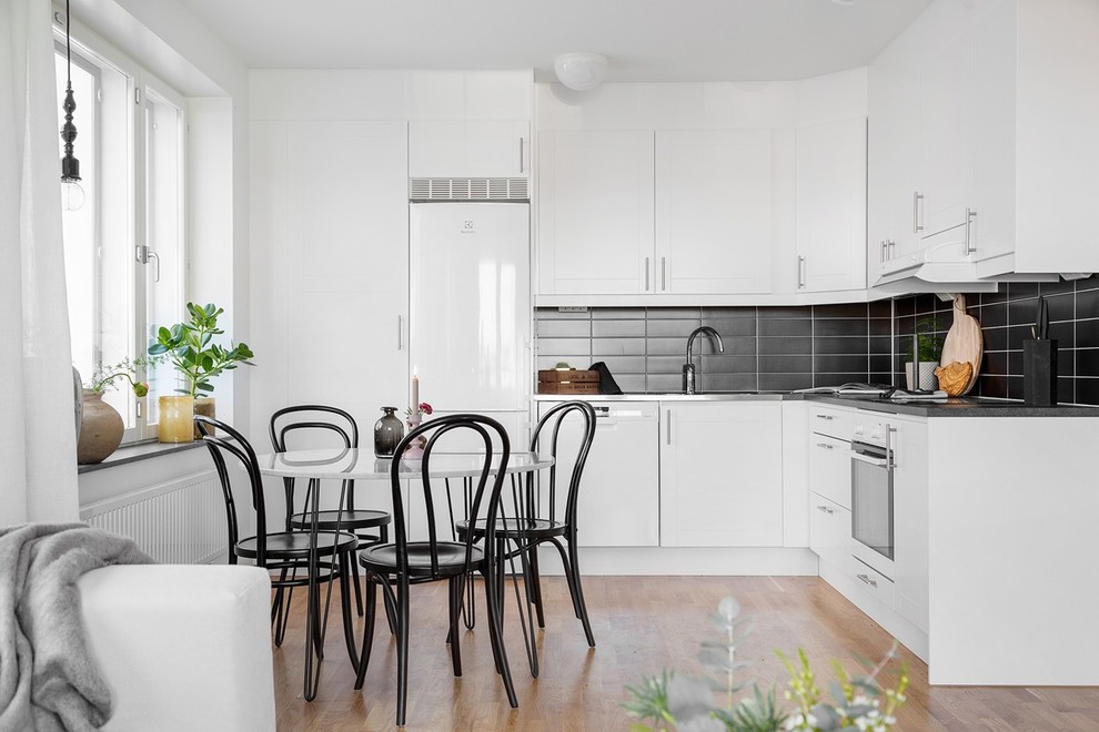 Photo of a scandinavian l-shaped eat-in kitchen in Stockholm with flat-panel cabinets, white cabinets, black splashback, white appliances, light hardwood floors, no island and brown floor.