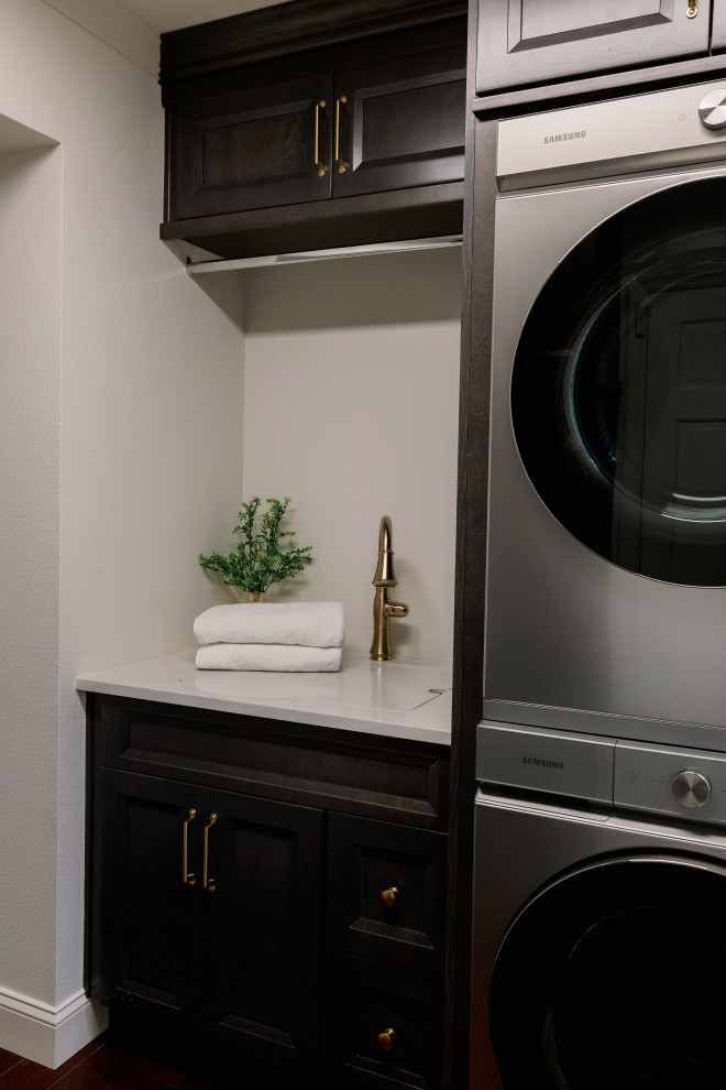 Mid-sized transitional single-wall dedicated laundry room in Orlando with a farmhouse sink, beaded inset cabinets, white cabinets, quartz benchtops, grey splashback, porcelain splashback, white walls, dark hardwood floors, a stacked washer and dryer, brown floor and grey benchtop.