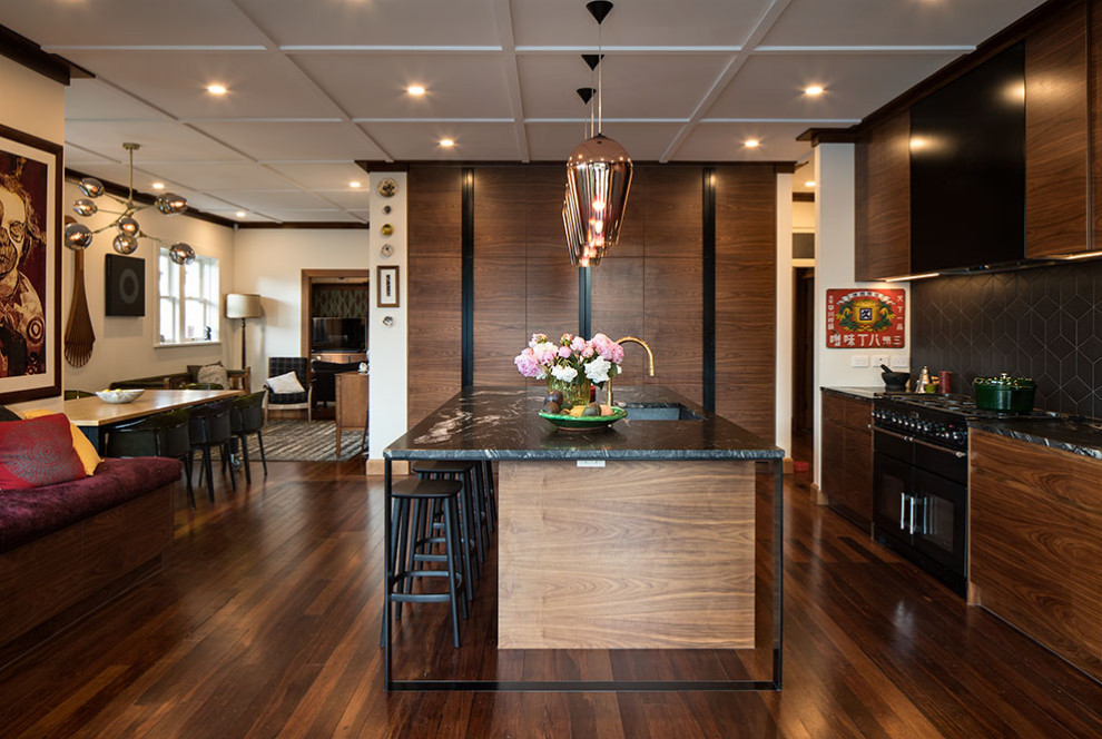This is an example of a large midcentury l-shaped eat-in kitchen in Auckland with a double-bowl sink, raised-panel cabinets, medium wood cabinets, granite benchtops, black splashback, ceramic splashback, black appliances, with island and black benchtop.