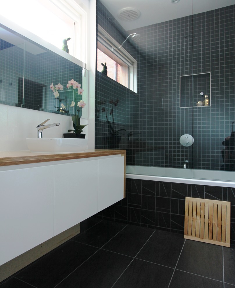 This is an example of a mid-sized contemporary master bathroom in Melbourne with white cabinets, wood benchtops, black tile, white walls, a shower/bathtub combo, a console sink and a two-piece toilet.