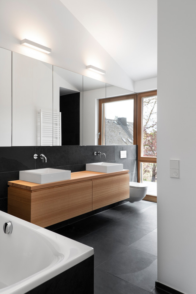 Photo of a large modern kids bathroom in Munich with flat-panel cabinets, light wood cabinets, a drop-in tub, a curbless shower, a wall-mount toilet, black tile, slate, white walls, slate floors, a vessel sink, wood benchtops, black floor, an open shower and brown benchtops.