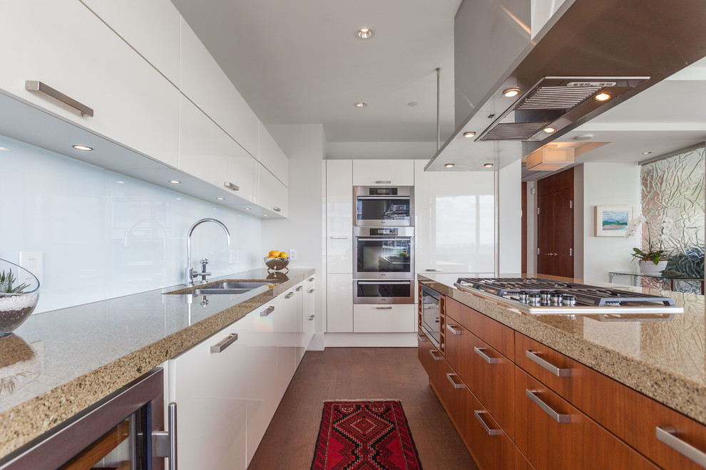 Mid-sized contemporary galley open plan kitchen in Vancouver with a double-bowl sink, flat-panel cabinets, white cabinets, white splashback, stainless steel appliances, medium hardwood floors, with island, brown floor and recycled glass benchtops.