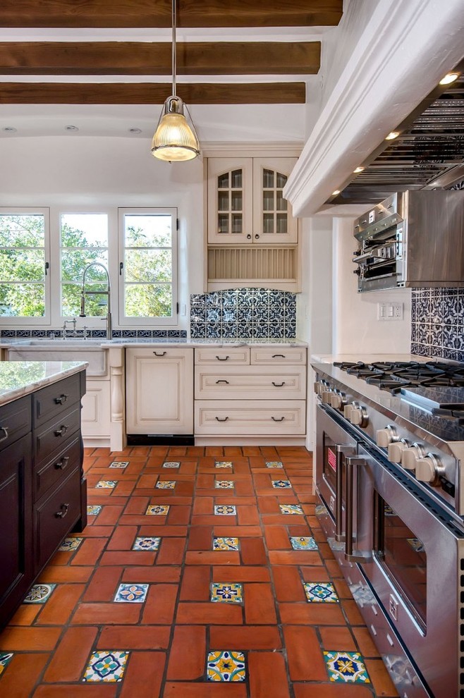 Photo of a mediterranean kitchen in San Francisco with a farmhouse sink, white cabinets, blue splashback, stainless steel appliances and terra-cotta floors.
