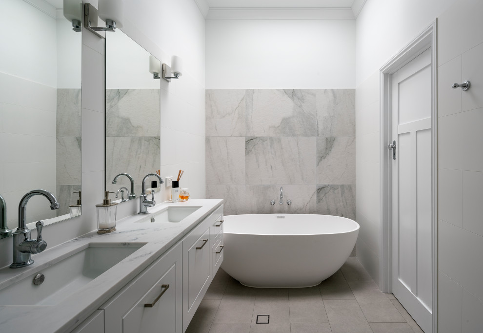 На фото: ванная комната в морском стиле с белыми фасадами, отдельно стоящей ванной, серой плиткой, белыми стенами, врезной раковиной, серым полом, белой столешницей, тумбой под две раковины и подвесной тумбой