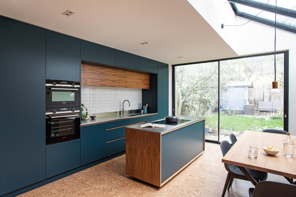 This is an example of a mid-sized contemporary eat-in kitchen in London with an integrated sink, flat-panel cabinets, blue cabinets, stainless steel benchtops, white splashback, cement tile splashback, black appliances, cork floors, with island and brown floor.