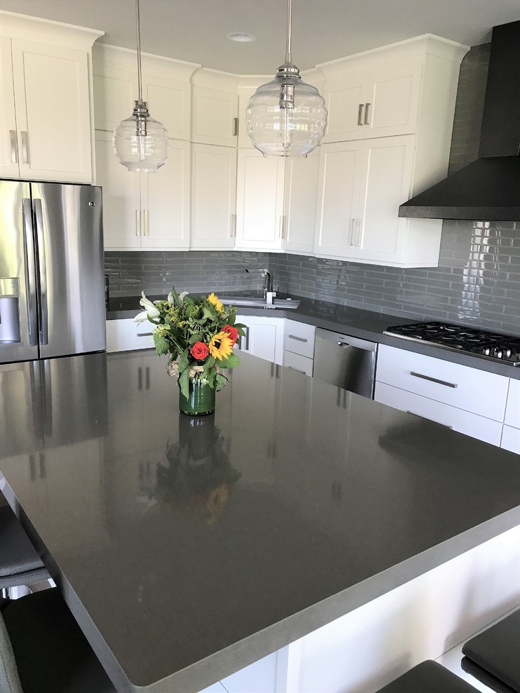 Mid-sized transitional l-shaped light wood floor and gray floor eat-in kitchen photo with an undermount sink, recessed-panel cabinets, white cabinets, quartz countertops, gray backsplash, ceramic backsplash, stainless steel appliances and an island