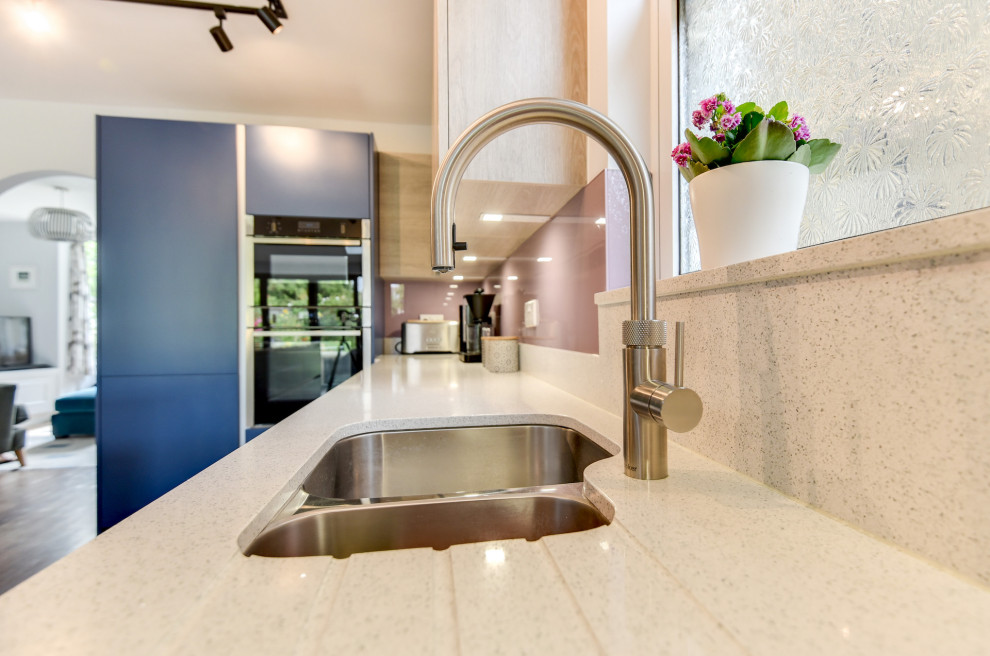Mid-sized beach style l-shaped eat-in kitchen in Sussex with a double-bowl sink, flat-panel cabinets, blue cabinets, quartzite benchtops, multi-coloured splashback, glass sheet splashback, black appliances, dark hardwood floors, with island, brown floor and white benchtop.