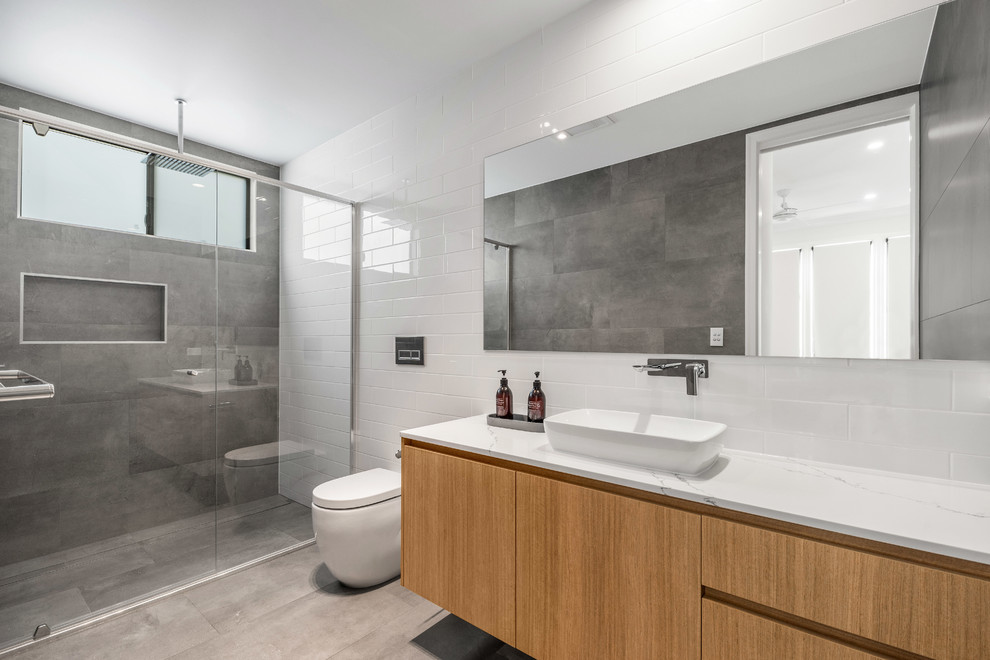 Photo of a mid-sized contemporary master bathroom in Brisbane with flat-panel cabinets, light wood cabinets, a freestanding tub, a corner shower, a one-piece toilet, gray tile, porcelain tile, grey walls, porcelain floors, a vessel sink, engineered quartz benchtops, grey floor and a hinged shower door.