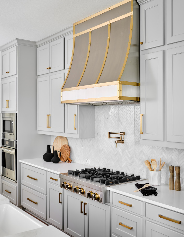 Belterra Project- Furnishings, Lighting Fixtures & Interior Design - This black, gray and gold urban farmhouse kitchen is the hub of the home for this busy family.  Our team changed out the existing plain kitchen hood for this showstopper custom stainless hood with gold strapping and rivets.  This provided a much needed focal point for this lovely kitchen.  In addition, we changed out the 36" refrigerator to a roomier 42" refrigerator and built-in a matching paneled refrigerator cabinet.  We also added the antique gold linear hardware and black and gold lighting to give it a streamlined look.  Touches of black tie the kitchen design into the rest of the home's mostly black and white color scheme.  The woven counter stools give the space a touch of casual elegance.  A new champagne gold kitchen faucet and potfiller add additional style, while greenery and wood accessories add a touch of warmth.