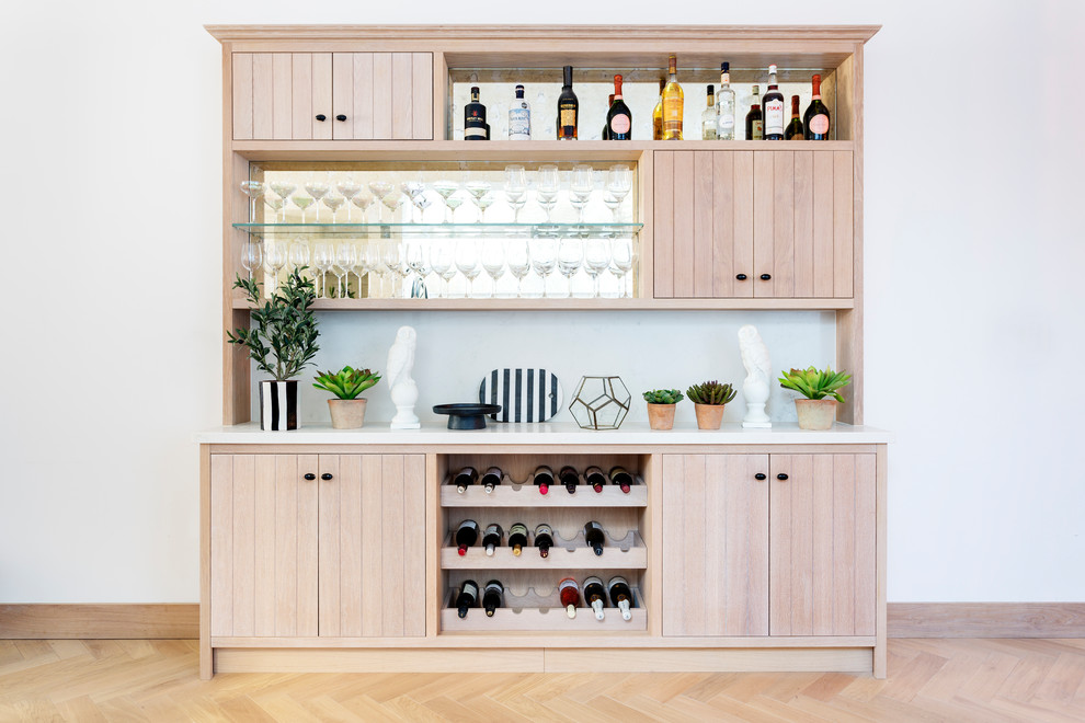 This is an example of a modern single-wall home bar in London with no sink, louvered cabinets, light wood cabinets, mirror splashback, light hardwood floors, beige floor and white benchtop.