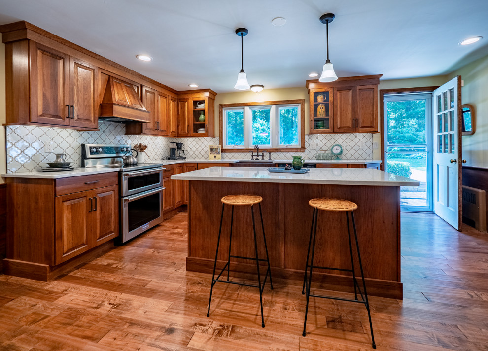 Design ideas for a mid-sized country l-shaped kitchen in Providence with a farmhouse sink, raised-panel cabinets, medium wood cabinets, quartz benchtops, beige splashback, ceramic splashback, stainless steel appliances, medium hardwood floors and with island.