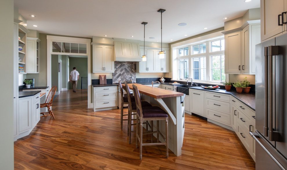 Photo of a transitional l-shaped kitchen in Burlington with a farmhouse sink, shaker cabinets, white cabinets, grey splashback, stainless steel appliances, medium hardwood floors, with island, brown floor and black benchtop.