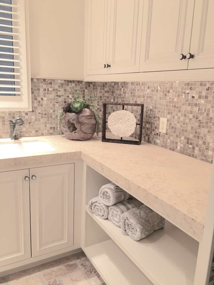 Design ideas for a mid-sized beach style u-shaped dedicated laundry room with raised-panel cabinets, white cabinets, limestone benchtops, white walls, limestone floors, a stacked washer and dryer and an undermount sink.