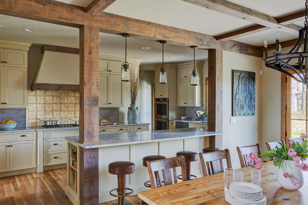 Example of a mid-sized mountain style u-shaped light wood floor, brown floor and exposed beam eat-in kitchen design in Chicago with a single-bowl sink, recessed-panel cabinets, beige cabinets, quartzite countertops, gray backsplash, cement tile backsplash, stainless steel appliances, a peninsula and gray countertops