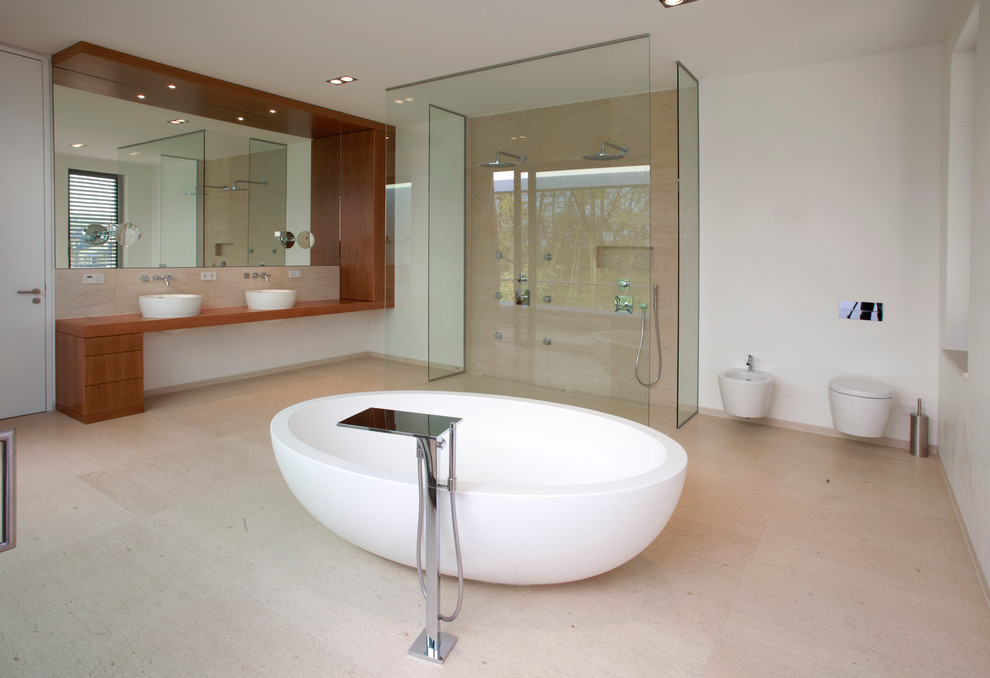 Photo of an expansive contemporary bathroom in Leipzig with flat-panel cabinets, dark wood cabinets, a freestanding tub, a curbless shower, a wall-mount toilet, beige tile, white walls, a vessel sink, wood benchtops, stone tile, limestone floors and brown benchtops.