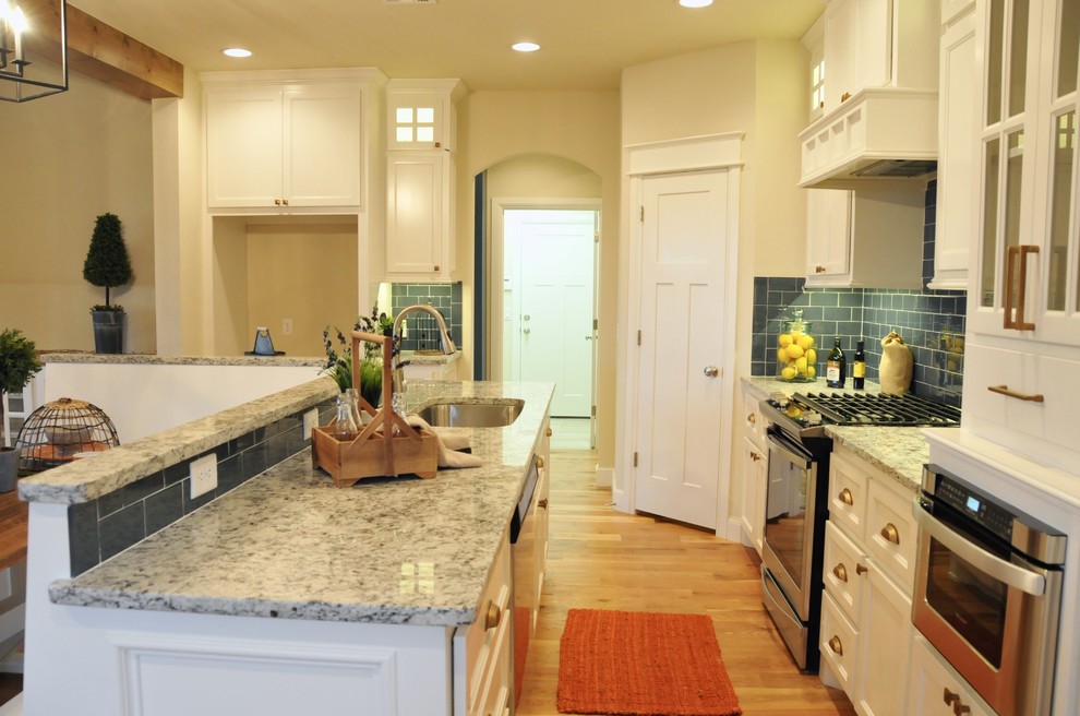 Design ideas for a mid-sized country l-shaped eat-in kitchen in Oklahoma City with an undermount sink, recessed-panel cabinets, white cabinets, granite benchtops, blue splashback, glass tile splashback, stainless steel appliances, light hardwood floors, with island and beige floor.
