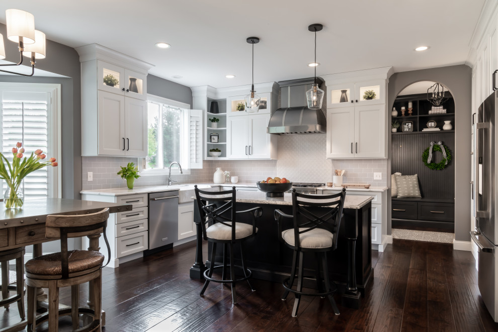 Example of a large transitional l-shaped dark wood floor and brown floor enclosed kitchen design in St Louis with an island, a farmhouse sink, shaker cabinets, white cabinets, quartz countertops, white backsplash, subway tile backsplash, stainless steel appliances and white countertops