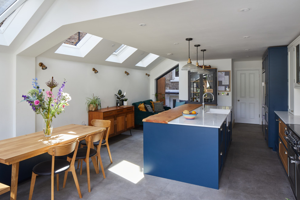 Photo of a medium sized traditional single-wall kitchen/diner in London with a belfast sink, shaker cabinets, blue cabinets, quartz worktops, white splashback, ceramic splashback, stainless steel appliances, porcelain flooring, an island, grey floors, grey worktops, a vaulted ceiling and feature lighting.