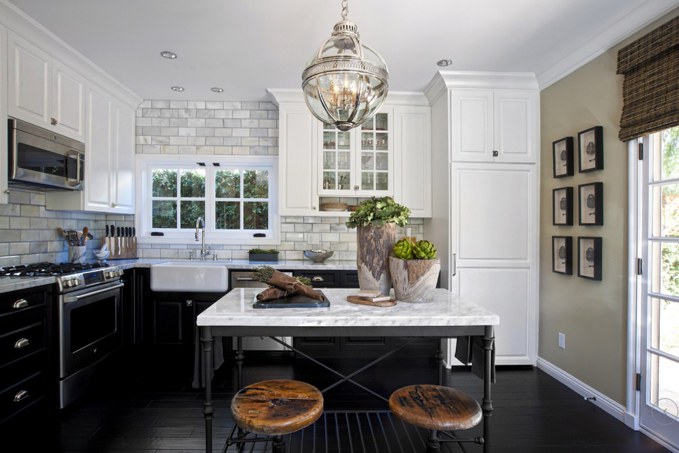 Photo of a mid-sized transitional l-shaped eat-in kitchen in Los Angeles with a farmhouse sink, raised-panel cabinets, black cabinets, marble benchtops, white splashback, subway tile splashback, stainless steel appliances, dark hardwood floors and with island.