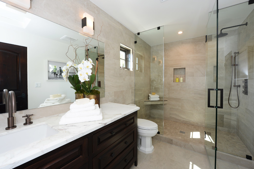 Inspiration for a mid-sized mediterranean master bathroom in Los Angeles with raised-panel cabinets, dark wood cabinets, a drop-in tub, an alcove shower, a one-piece toilet, white tile, travertine, beige walls, travertine floors, an undermount sink, marble benchtops, beige floor, a hinged shower door and white benchtops.