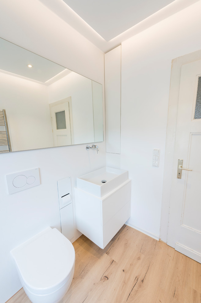 This is an example of a mid-sized contemporary 3/4 bathroom in Other with white cabinets, a curbless shower, a wall-mount toilet, white walls, medium hardwood floors, a vessel sink, brown floor, an open shower, white benchtops, flat-panel cabinets, black and white tile, pebble tile and solid surface benchtops.