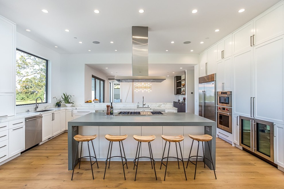 Contemporary u-shaped kitchen in San Francisco with an undermount sink, shaker cabinets, white cabinets, marble benchtops, white splashback, stone slab splashback, stainless steel appliances, medium hardwood floors, with island and brown floor.