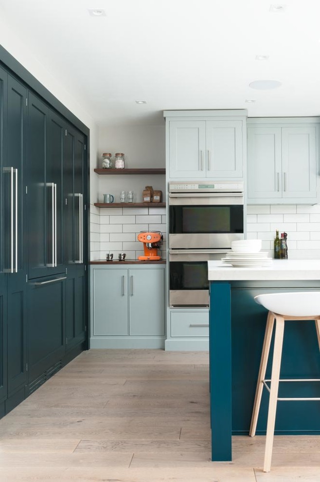 Inspiration for a large contemporary single-wall open plan kitchen in Kent with a drop-in sink, recessed-panel cabinets, blue cabinets, solid surface benchtops, white splashback, ceramic splashback, stainless steel appliances, light hardwood floors and with island.