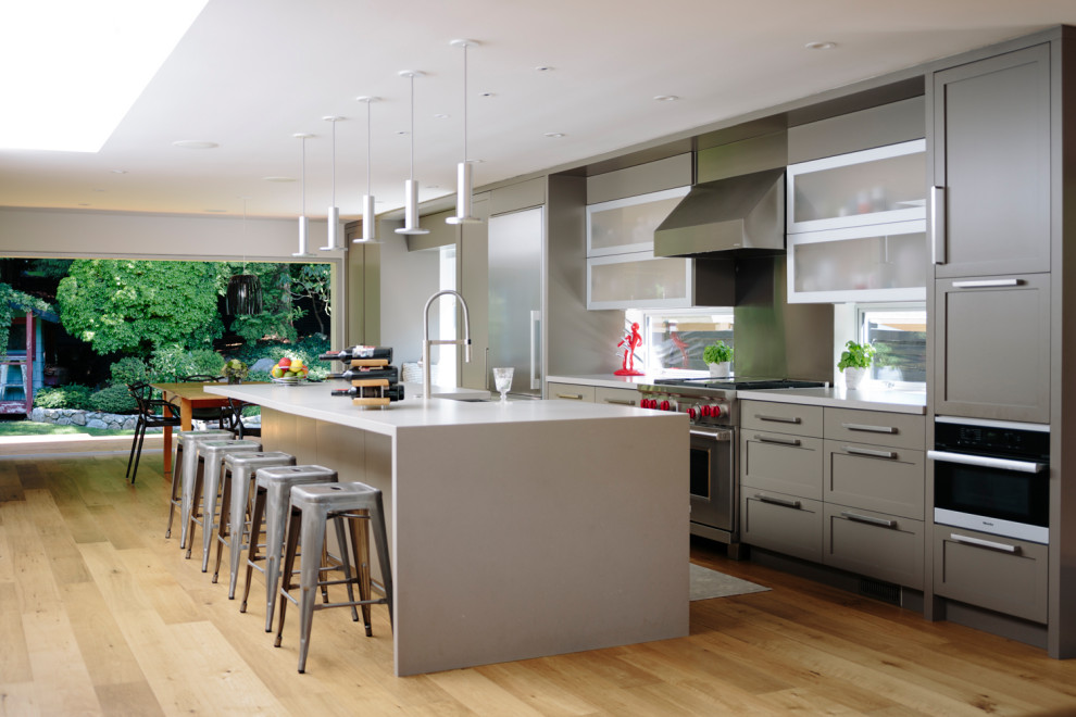 Photo of a contemporary kitchen in Vancouver with shaker cabinets, grey cabinets, stainless steel appliances, light hardwood floors, with island and white benchtop.