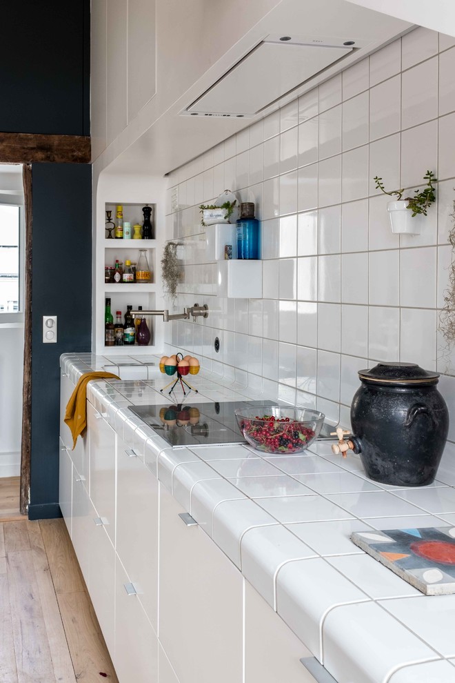 This is an example of a mid-sized contemporary single-wall open plan kitchen in Paris with flat-panel cabinets, white cabinets, tile benchtops, white splashback, porcelain splashback, panelled appliances, light hardwood floors, beige floor, white benchtop, an undermount sink and no island.