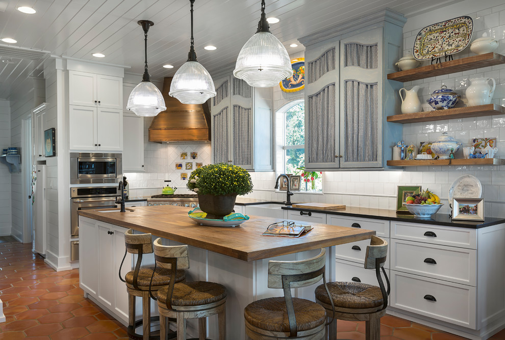 Beach style l-shaped kitchen in Charlotte with a farmhouse sink, recessed-panel cabinets, white cabinets, white splashback, stainless steel appliances, terra-cotta floors, with island, orange floor and black benchtop.