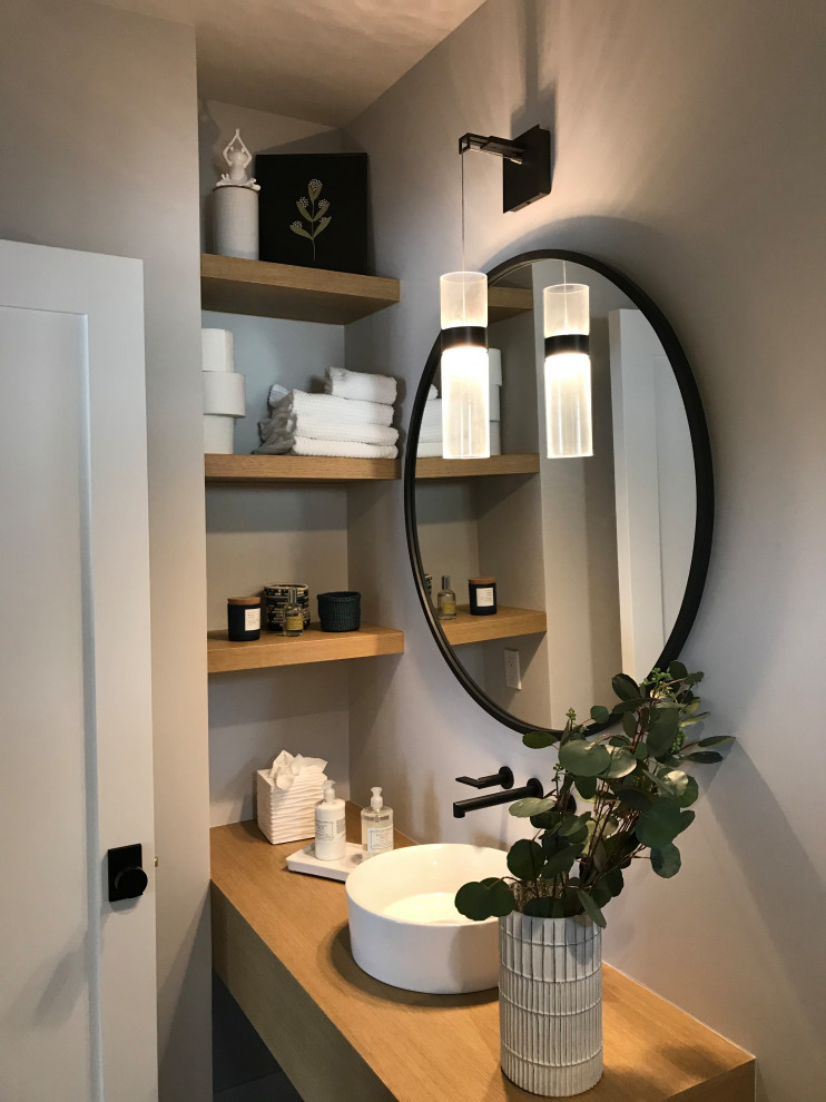 Foto på ett toalett, med skåp i ljust trä, grå väggar, ett fristående handfat och träbänkskiva