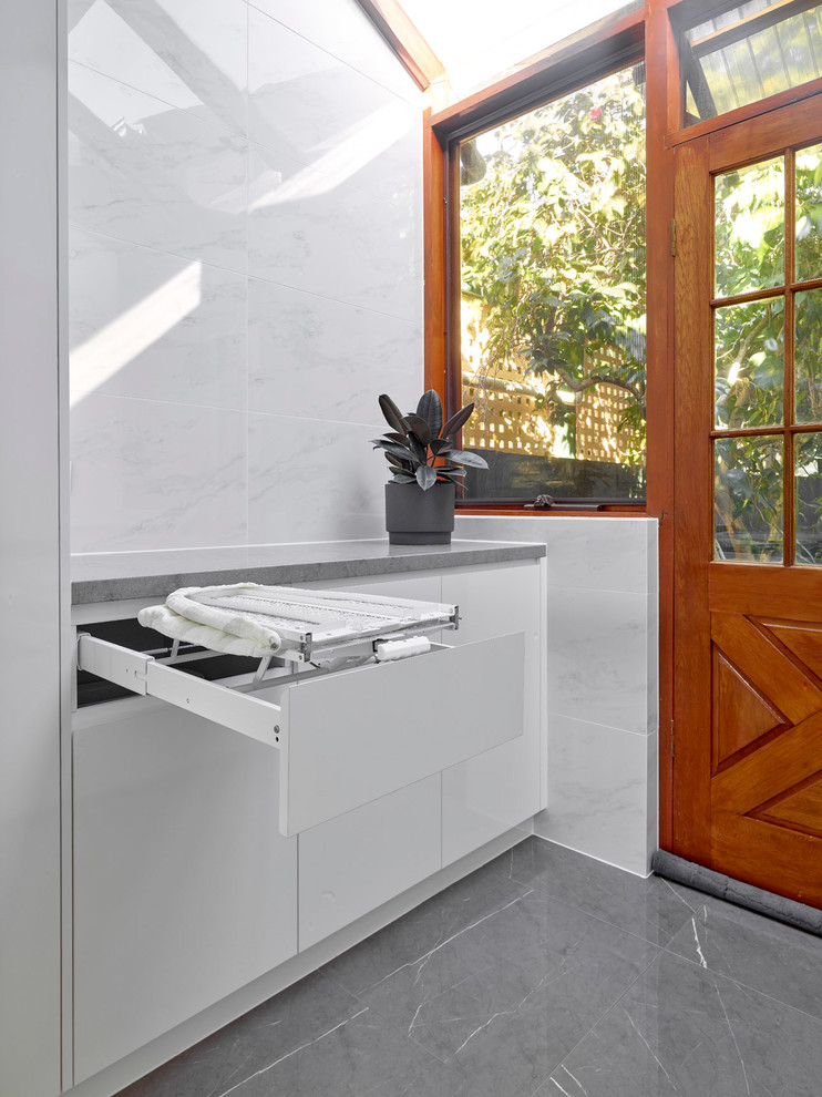 Mid-sized modern galley dedicated laundry room in Other with a drop-in sink, flat-panel cabinets, white cabinets, quartz benchtops, white walls, porcelain floors, a concealed washer and dryer, grey floor and grey benchtop.