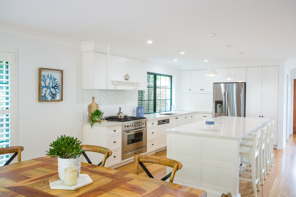 Inspiration for an expansive traditional galley eat-in kitchen in Brisbane with a single-bowl sink, shaker cabinets, white cabinets, solid surface benchtops, white splashback, subway tile splashback, stainless steel appliances, medium hardwood floors, with island, brown floor and white benchtop.