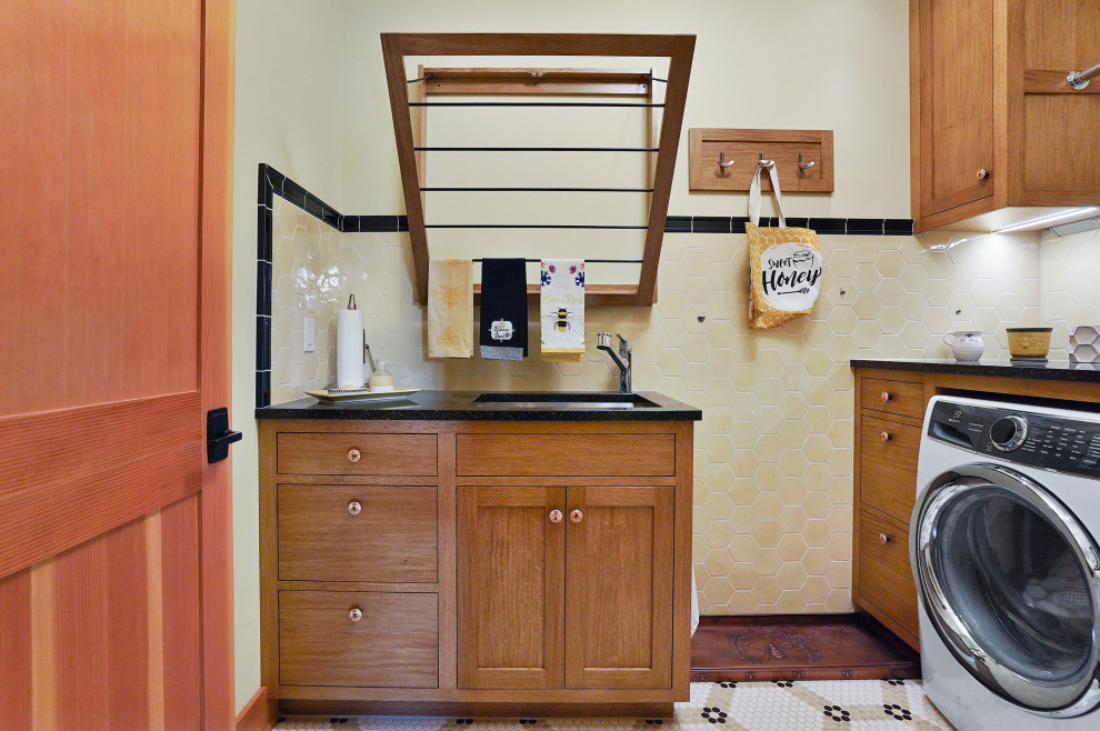 Laundry room - large craftsman porcelain tile and wainscoting laundry room idea in Other with an utility sink, shaker cabinets, medium tone wood cabinets, granite countertops, yellow backsplash, ceramic backsplash, yellow walls, a side-by-side washer/dryer and black countertops