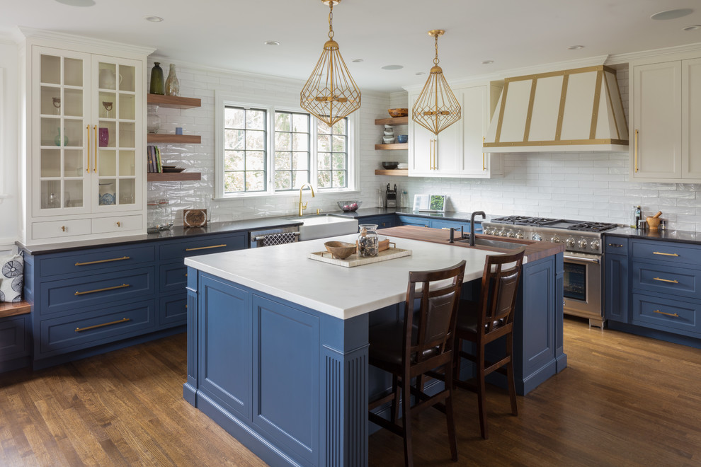 Photo of a traditional l-shaped kitchen in New York with a farmhouse sink, recessed-panel cabinets, blue cabinets, white splashback, subway tile splashback, stainless steel appliances, medium hardwood floors, with island and brown floor.