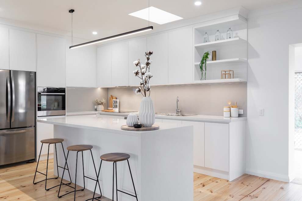 Design ideas for a contemporary l-shaped kitchen in Adelaide with a drop-in sink, flat-panel cabinets, white cabinets, quartz benchtops, grey splashback, glass sheet splashback, stainless steel appliances, light hardwood floors, with island, white benchtop and beige floor.