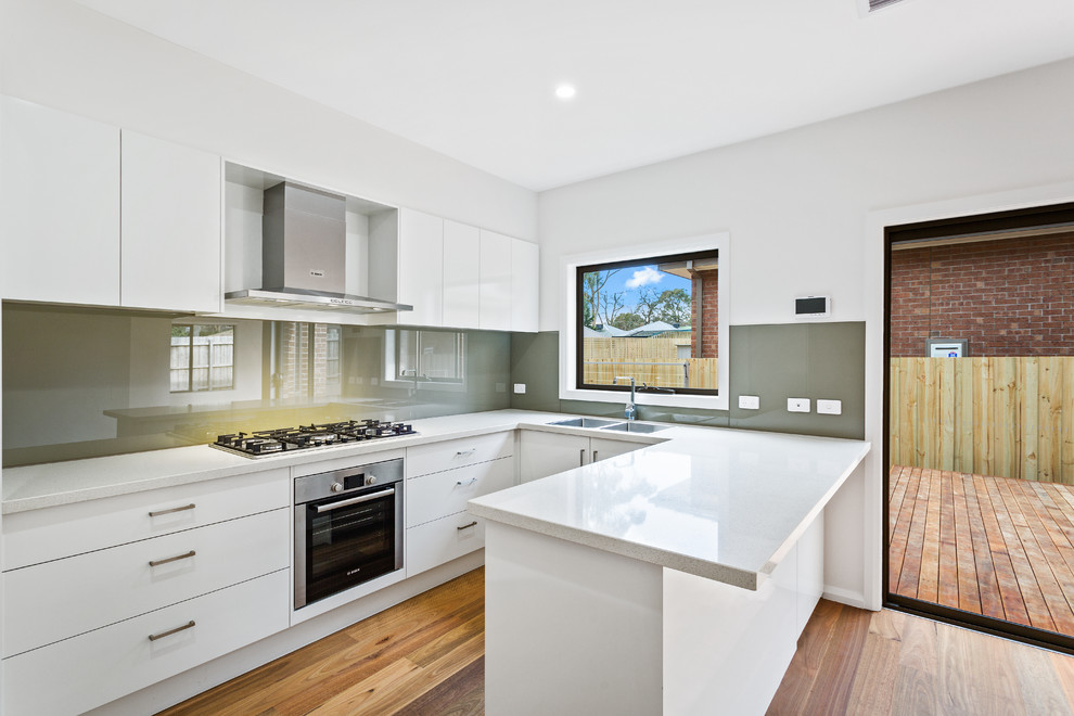 Photo of a mid-sized modern u-shaped separate kitchen in Melbourne with a drop-in sink, flat-panel cabinets, white cabinets, quartz benchtops, brown splashback, glass sheet splashback, stainless steel appliances, medium hardwood floors, no island and brown floor.