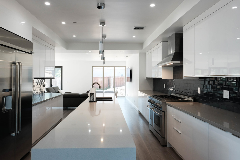 Large modern galley eat-in kitchen in Los Angeles with an integrated sink, flat-panel cabinets, white cabinets, quartz benchtops, black splashback, ceramic splashback, stainless steel appliances, medium hardwood floors, with island and brown floor.