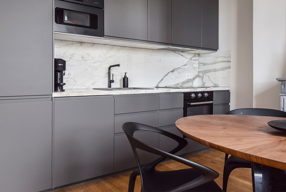 Photo of a medium sized classic grey and white single-wall open plan kitchen in Paris with a submerged sink, flat-panel cabinets, grey cabinets, marble worktops, white splashback, marble splashback, black appliances, light hardwood flooring, no island, beige floors and white worktops.