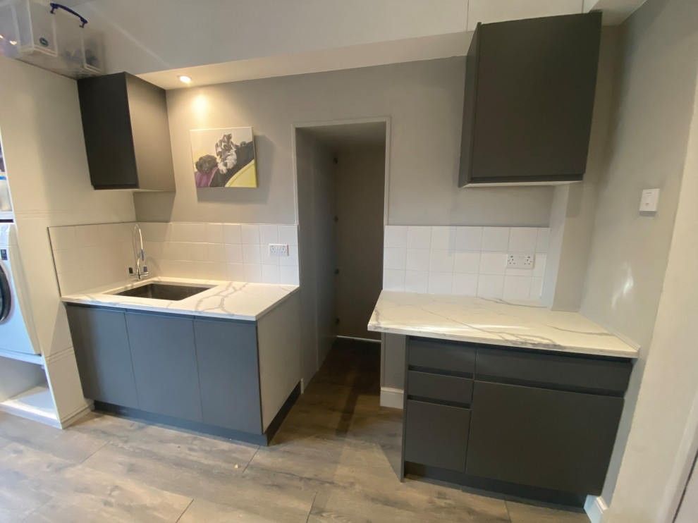Photo of a large contemporary galley utility room in London with a drop-in sink, flat-panel cabinets, grey cabinets, quartzite benchtops, white splashback, porcelain splashback, beige walls, laminate floors, a side-by-side washer and dryer, grey floor and multi-coloured benchtop.