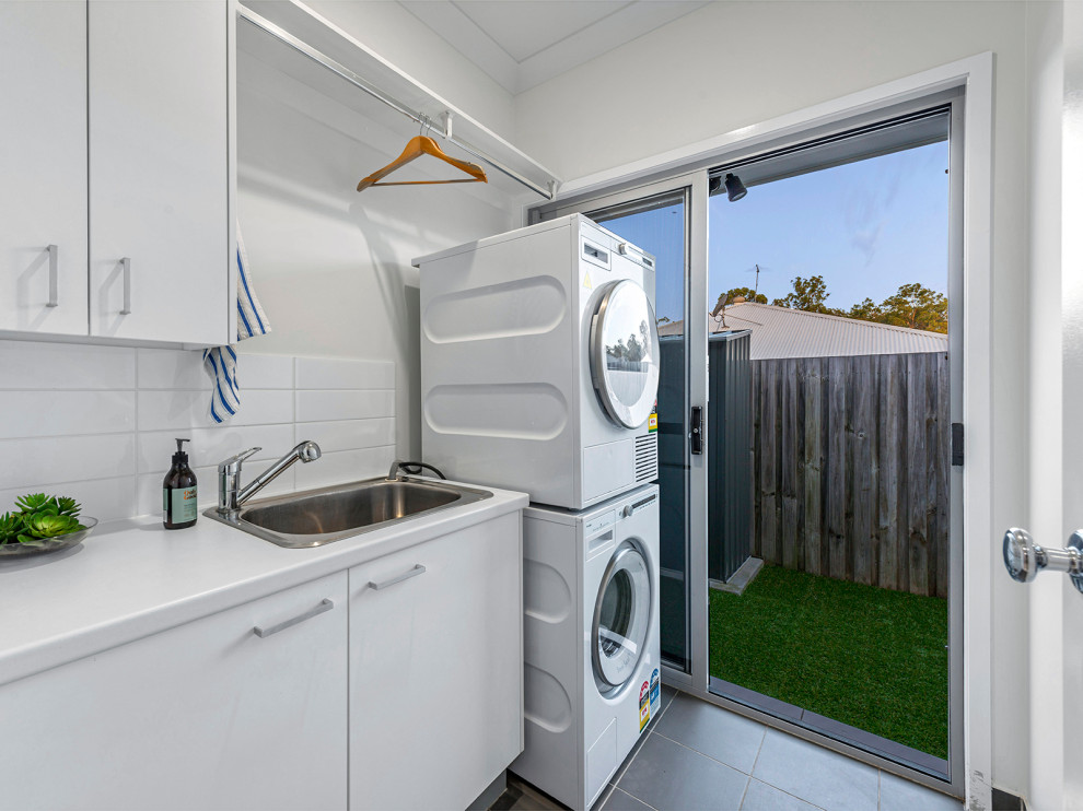 Small beach style single-wall laundry room in Brisbane with a drop-in sink, flat-panel cabinets, white cabinets, laminate benchtops, white walls, ceramic floors, a stacked washer and dryer, grey floor and white benchtop.