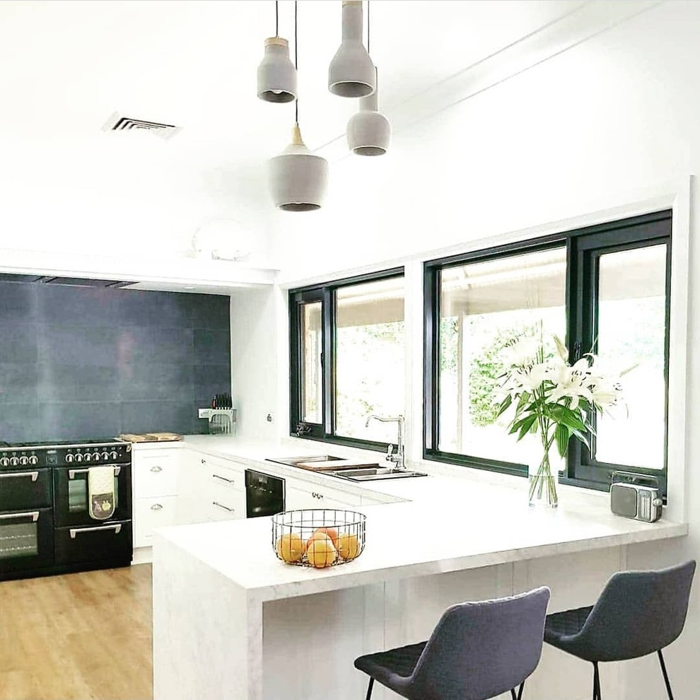Photo of a mid-sized contemporary u-shaped kitchen pantry with a drop-in sink, raised-panel cabinets, white cabinets, quartz benchtops, grey splashback, ceramic splashback, black appliances, medium hardwood floors, with island, brown floor and white benchtop.