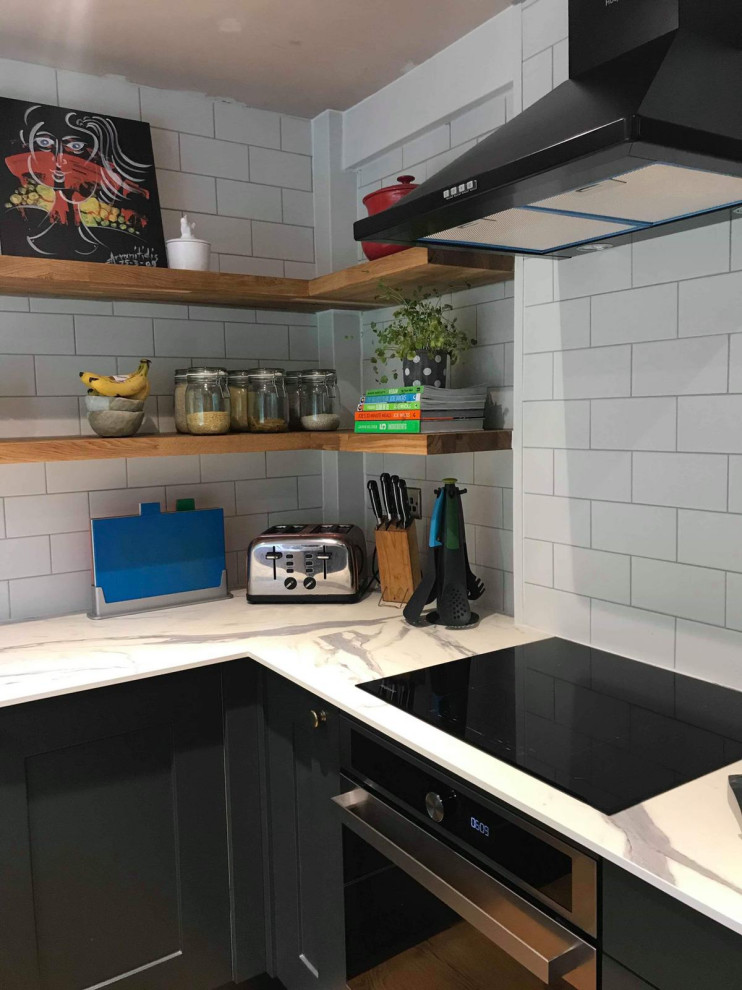 Photo of a small traditional galley enclosed kitchen in Other with a belfast sink, shaker cabinets, green cabinets, quartz worktops, white splashback, brick splashback, black appliances, light hardwood flooring, brown floors, white worktops and a chimney breast.